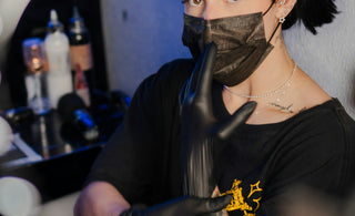 Tattooed woman in black mask and gloves making shush gesture.