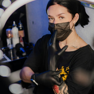 Tattooed woman in black mask and gloves making shush gesture.