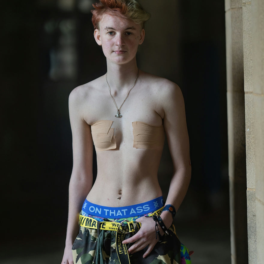 A person with short dyed hair wearing chest tape and camouflage trousers